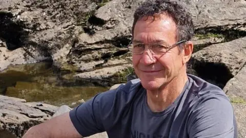 David Goodman, with short brown hair, wearing a navy T-shirt and sitting on rocks with a small pool of water behind.