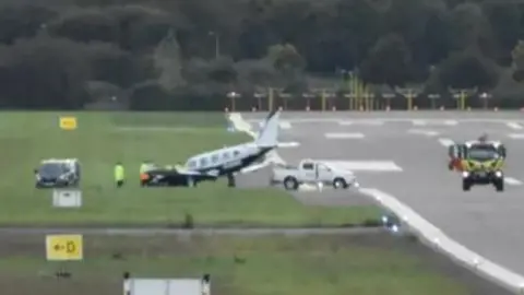 A light aircraft on a field next to a runway surrounded by cars