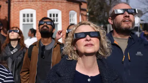 Neil Hall/Shutterstock A group of people are standing in front of a red brick observatory. They are all wearing eclipse glasses with dark lenses and black cardboard frames.