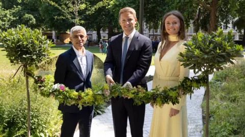 Maddie Cook Sadiq Khan, Hugh Grosvenor (the Duke of Westminster), and Olivia Grosvenor (the Duchess of Westminster) standing side-by-side on a paved pathway, holding a long garland made of green foliage and small flowers. Sadiq Khan on the left and Hugh Grosvenor in the middle wear dark suits and collars, while Olivia Grosvenor on the right wears a light-coloured dress. They are positioned between two small, pruned potted trees, with a park lawn and trees in the background.