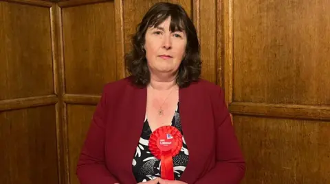 Isabella Harris Susan Brown, standing next to a wood-paneled wall in Oxford Town Hall, is wearing a red jacket and bright red rosette. She has long dark hair.