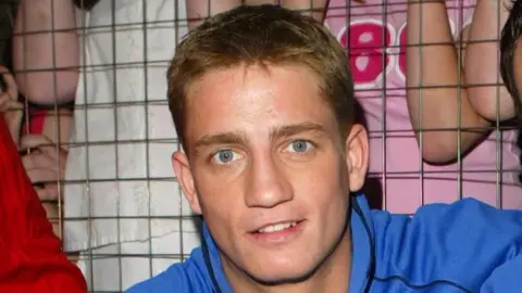 Getty Philip Olivier is pictured during his Brookside years while on SMTV Live. He is wearing a blue hooded jumper with the words "Brookside team". He has short blonde hair and is smiling at the camera. 