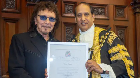 Iommi is wearing black with his trademark tinted glasses and long curly hair and beard. Iqbal is in mayrol finery, which is gold and black with large chain around his neck. He is holding the award to give to Iommi. Iqbal has short dark hair.