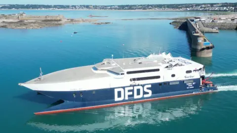 A white and blue ferry with the logo DFDS in white along the side is sailing in St Helier. It is s bright sunny day and the water is turquoise.
