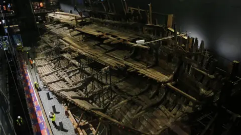Getty Images The remains of a large wooden ship are pictured on a stage, with several people in hi-viz vests walking around its base.
