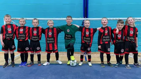 Young girls and boys standing in a row, in a sports hall. They are in front of a football goal and have their arms around each other's shoulders. They are all wearing a red and black striped football kit. 