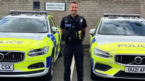 Cambridgeshire Constabulary PC Boddington has short light brown hair and is standing in his uniform and smiling. He is standing between two marked police cars and against a wall. There is a sign in the background that says "Marked Police Vehicles Only".