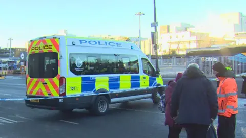 BBC A police van is stationed across the lanes of a road outside a bus station. Police tape blocks the road. 