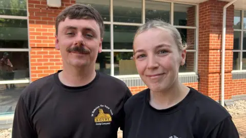 Lance Corporal Daniel Stapley & Corporal Chloe Stapley both smile at the camera. They're wearing 1st Battalion the Royal Welsh black t-shirts. Daniel has a moustache. Chloe's blonde hair is tied back. 