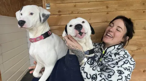 Two white Dalmatian dogs on the knee of a woman. The woman has dark hair and is wearing a black and white zipped jumper. She is smiling and giving the dogs plenty of attention.