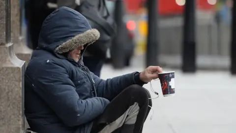 A homeless man wearing a hood holds out a paper cup.