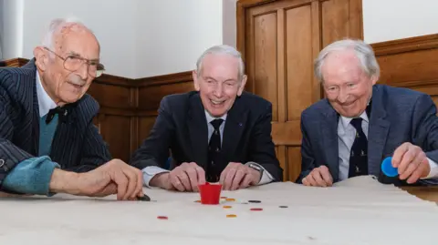 Bill Steen, Peter Downes and Lawford Howells are sat together at a table with a white mat on top. All three men are smiling as they play a game of tiddlywinks. There a different coloured counters atop the table as well as a small red cup. Mr Steen wears large golden framed glasses, a striped blazer, grey jumper and black bow tie. He has grey hair and is reaching for a counter on the table. Mr Downes also has grey hair and wears a black blazer, watch on his wrist, tie and white shirt. Mr Howells has grey hair, a blue blazer, tie, white shirt and is holding a large blue counter in his hand. 