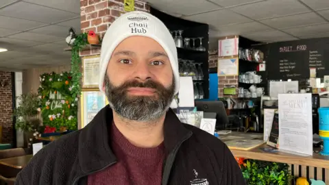Tom Jackson/BBC Sunny Mann is standing off centre to the left. He has a short brown and grey beard, and is wearing a white hat which says Chaii Hub on it, a burgundy jumper and black jacket. He is standing in front of a bar inside.