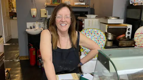 Jonny Humphries/BBC Caroline Ashton, in her 50ss who has long, straight brown hair and wears a black top under a black apron, smiles at the camera from behind the counter of a cafe