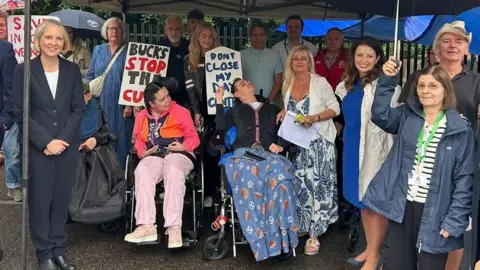 Hazel Howe Group of people gathered under a canopy and umbrellas at a protest, including wheelchair users at the front, holding signs reading “Save Burnham Day Centre”, “Bucks Stop the Cuts” and “Don’t Close My Centre”.