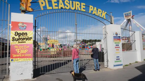 An older couple with a dog peer through closed gates at a 'tea cups' ride, 'Pleasure Park' is written above the gates.