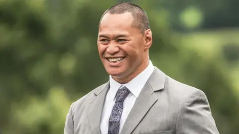 A man with short dark hair wearing a purple patterned tie, white shirt and light grey suit jacket smiles while looking to his right. 