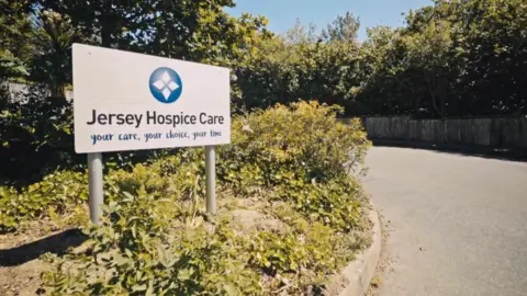 BBC A white sign with black and blue writing reading 'Jersey Hospice Care - your care, your choice, your time' and a round blue circle logo with four diamonds inside. The sign is in a bed of foliage and is next to a Tarmac driveway. More foliage is visible in the background. 