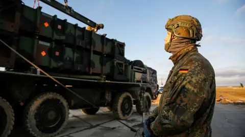 A soldier with a German flag patch sewn into the arm of their uniform operates a remote while looking up at a large military truck moving along a road against a blue sky, as German military moved air defence units to southeast Poland in January.