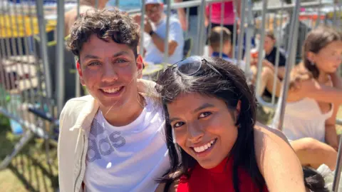 Alice Cunningham/BBC Reuben Librack and Senumi Tebuwna smile at the camera while sitting in a queue outside Portman Road. Reuben has dark curly hair and Senumi has long dark hair.