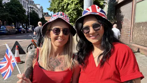 Two women smiling, in red dresses. They are holding red, white and blue Union Jacks and wearing union jack hats