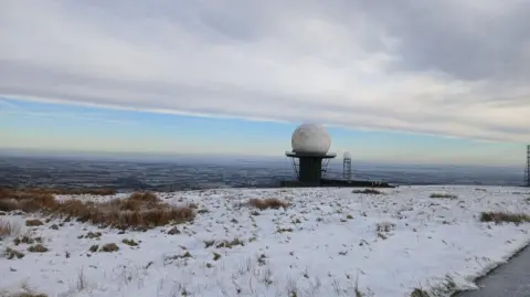 Jane Watkins A huge golf-ball like radar on top of a large platform with a fence around its perimeter. The dome is white and has hexagonal shapes on its outside. In the background there is a smaller radar of the same shape, also on a tower. Grass around it is covered in snow. In the distance are hills, towns and fields. The sky is blue with grey and white clouds