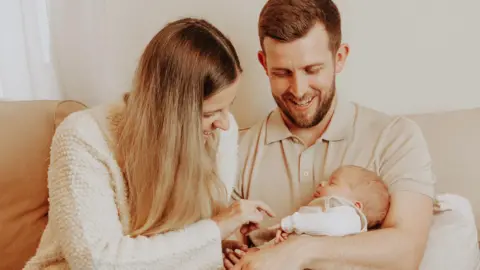 Beth Kelly A professional looking shot of a couple, Beth Kelly and Ben Stanbrook, sat on a sofa with their baby son, Wilfred. Both Kelly and Stanbrook are looking down at Wilfred and smiling.