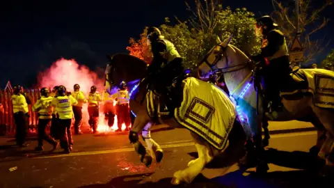 Police horses and riders in the foreground are among a group of police officers who are tackling disorder. It is night time, and the officers on foot are tackling what seems to be a flare. One of the horses is rearing up on its back legs. They are wearing yellow high visibility covers.