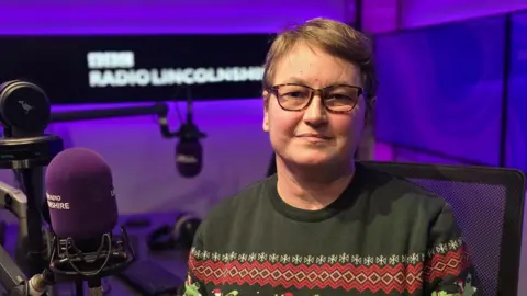 Head and shoulders shot of Maggie Cullen seated in the BBC Radio Lincolnshire studio. She is wearing spectacles and a green and red Christmas jumper.