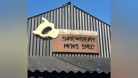 Simon Rouse A corrugated metal building with a large brown sign on it. The sign reads "Shrewsbury Men's Shed". There is blue sky in the background