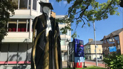 Orla Moore/BBC A picture of the Cambridge Don statue. It depicts a white statue wearing a university robe and hat in black and gold. It sits in front of a building near trees. Other buildings and a road can be seen in the background.