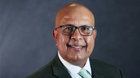 A picture of Sajjad Malik, who is wearing a black jacket, green tie and white shirt. He is smiling and is wearing glasses. He is sat or stood against a grey background.