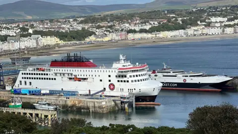 The Manxman and Manannan ferries, which are both painted in the Isle of Man Steam Packet Company's colours of white, red and black, moored in Douglas Harbour. Douglas Bay and the tall white buildings on Douglas Promenade can be seen in the background.