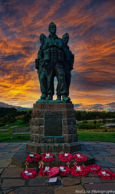 Neil Lea A bronze war memorial statue stands tall on a stone pedestal inscribed with the words “united we conquer.” The base is adorned with red poppy wreaths and wooden crosses, symbolising remembrance and respect.
Behind the monument, a dramatic sunset fills the sky with fiery orange and deep purple hues.