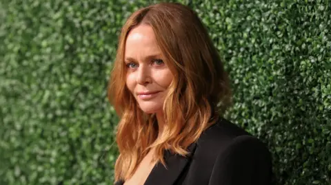 Stella McCartney has long blonde hair and is wearing a black suit. She is standing next to a green hedge.