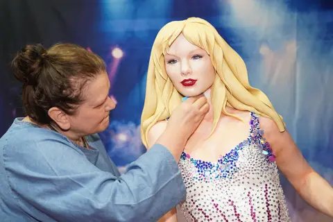 Jacob King/PA Media Cakemaker Elza Baldzhiyska putting finishing touches to her Taylor Swift cake. Wearing a light blue top, Ms Baldzhiyska is smoothing icing around the neck of her creation, with Swift depicted in a sparkling outfit from her Eras tour