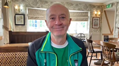 A man wearting a green T-shirt under a light green and black jacket sat inside a room smiling softly at the camera