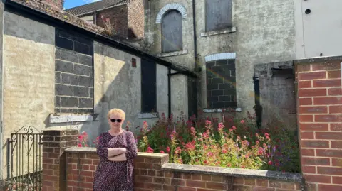BBC Kathleen Wilson is stood with her arms crossed outside a very overgrown backyard. The house behind it has cracked paint and all of the windows have been boarded up or filled with concrete blocks. 
