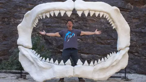 Louis Smith/Daniel Southwell Steve Backshall, wearing a navy blue T-shirt with shark drawings printed on it, is standing inside a giant shark jaw replica or sculpture. Surrounding him is a massive circular jaw structure with large, pointed teeth on both the upper and lower jaws.