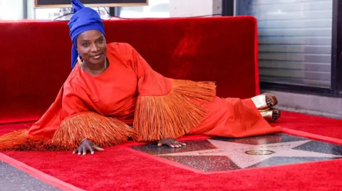Angélique Kidjo lying down on a red carpet behind her star which is on the ground. She is wearing a red, long-sleeved dress with tasselled sleeves and a royal blue headwrap