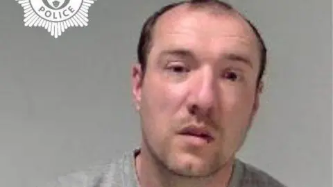 A man with black receding hair is photographed in a grey sweatshirt in front of a grey background. 