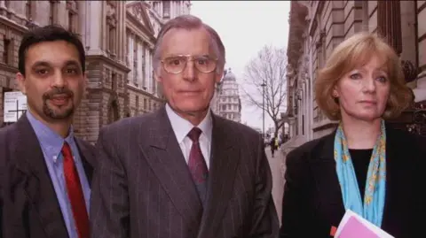 PA Media Lord Laming (centre) with Prof Hazel Genn (right) and Dr Aneez Esmail (left) poses for a photo taken in 2000 in a street in Westminster, London.