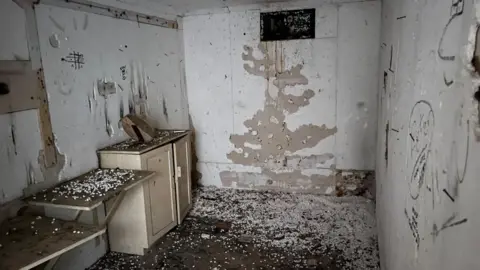 Andrew Turner/BBC The bunker interior, with deteriorated polystyrene insulation on the walls, with graffiti, debris on the floor and a cupboard and shelves on the left and a ventilation shaft central on the upper part of the back wall