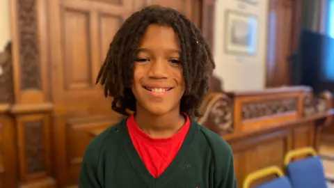 Ethan has brown hair that sits on top of his shoulders. He is wearing school uniform; a red t-shirt under a green v-neck jumper. He is in an old hall with brown wooden panelling, and there are two blue chair behind him.