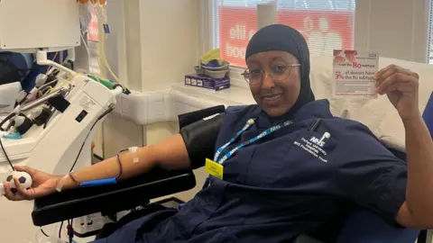 NHS Blood and Transplant Ms Ali, still wearing glasses, a head covering and a dark blue nurse uniform,   smiles as she donates blood in the chair. She has a needle in her right arm, which also hold a football stress ball, and holds up a donation card to the camera with her other hand.