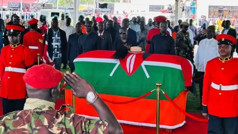 An officer in a red beret seen from behind as he salutes at the coffin of Raila Odinga, which is draped in the Kenya flag on which sits a cream hat and fly whisk. Officials stand behind it.