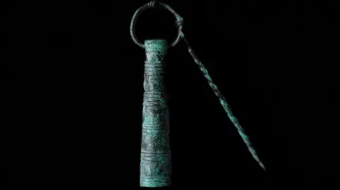 Discover Bucks Museum An early Saxon bronze toiletry set which consists of a hollow cylinder, slightly narrower at the top and wider at the bottom. There is a ring through its top end and attached to it is a twisted bronze rod. The bronze is heavily flecked in green oxidisation.