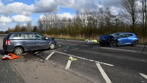 An otherwise empty main road with a grey car on the left and a blue car on the right, facing each other across the carriageway. Both cars have their fronts smashed in and there is debris across the road.