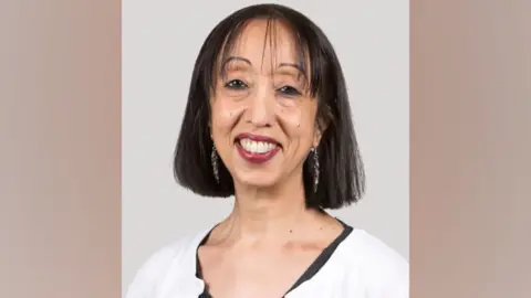 Hodge Jones & Allen Solicitors Nina Ali has straight dark hair down to her neckline. She is smiling at the camera and has dark red lipstick on. She is wearing a white cardigan with black top underneath, and has long earrings on as well.