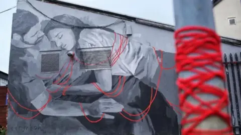 Caryn Koh A black and white mural of a man and a woman in a cuddling position entangled in red threads. In the foreground red thread is wrapped around a street post and appears to be connected to the red threads on the mural.